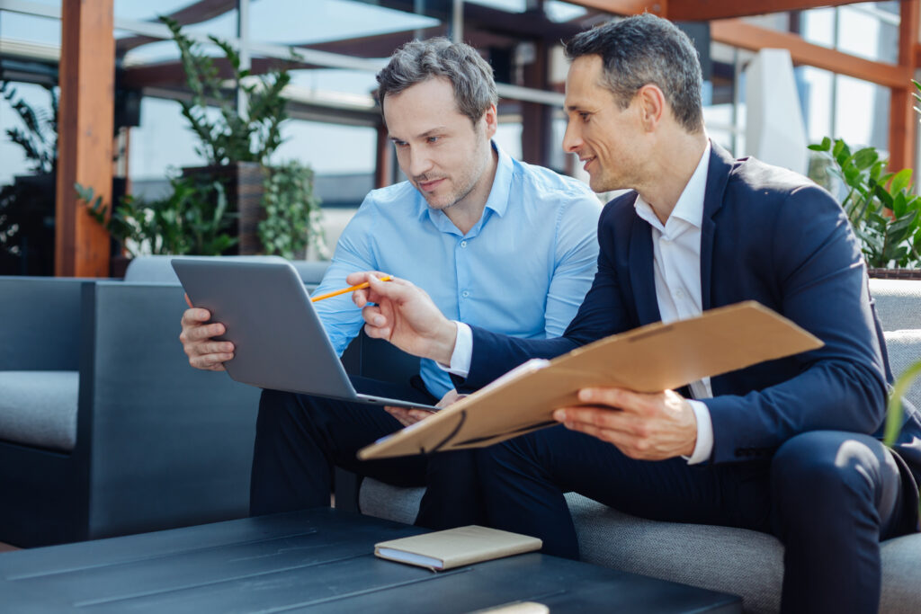 Two businessmen with a laptop and folder of documents discussing changes to financial statement assertions