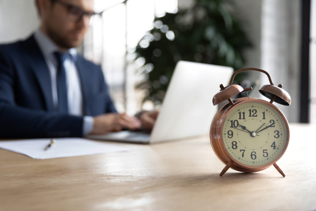 Close up of clock on forefront with businessman working on ERISA audit requirements on his laptop in office in background