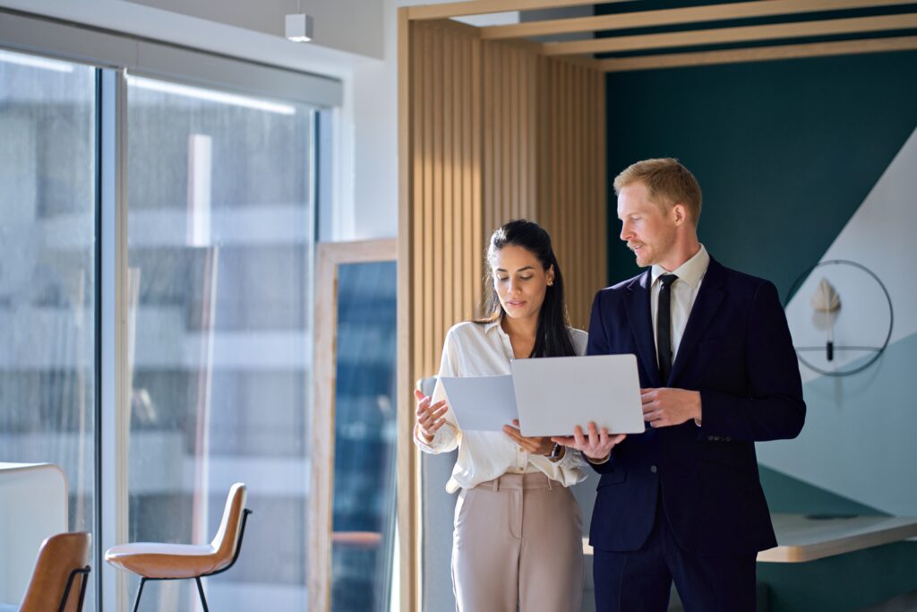 Two business executives reviewing a public company audit report on a laptop in an office hallway