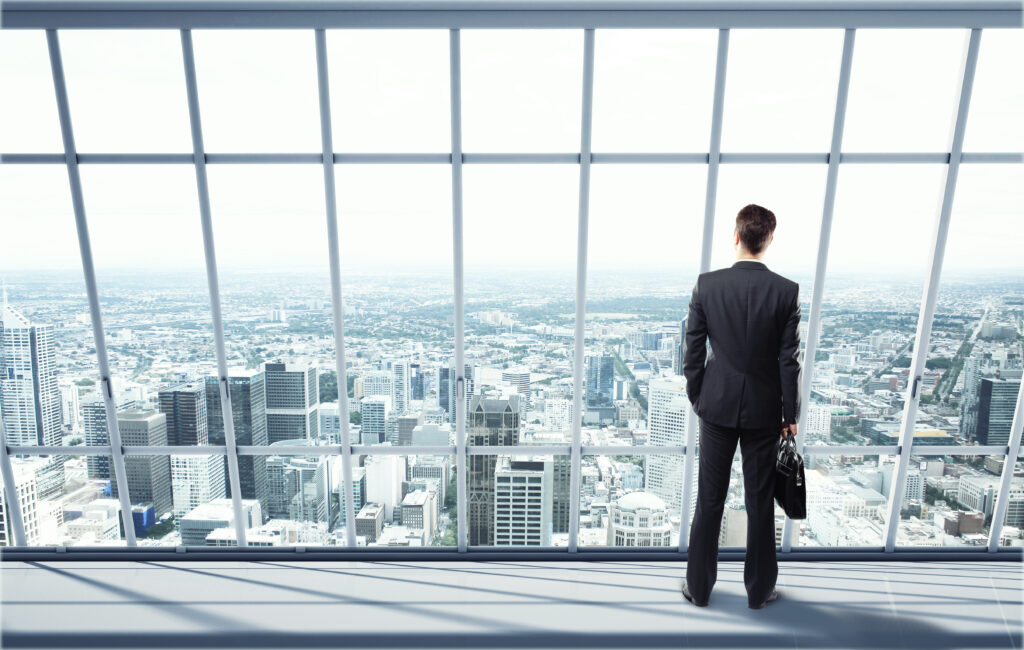 A man in a business suit looking out the window at a city wondering "public accounting firms are near me" 