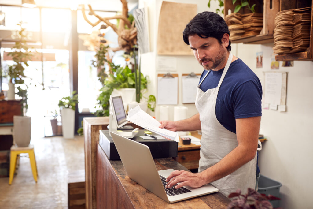 Man in an apron reviewing documents and working on a laptop, researching paperwork from his franchise auditor.