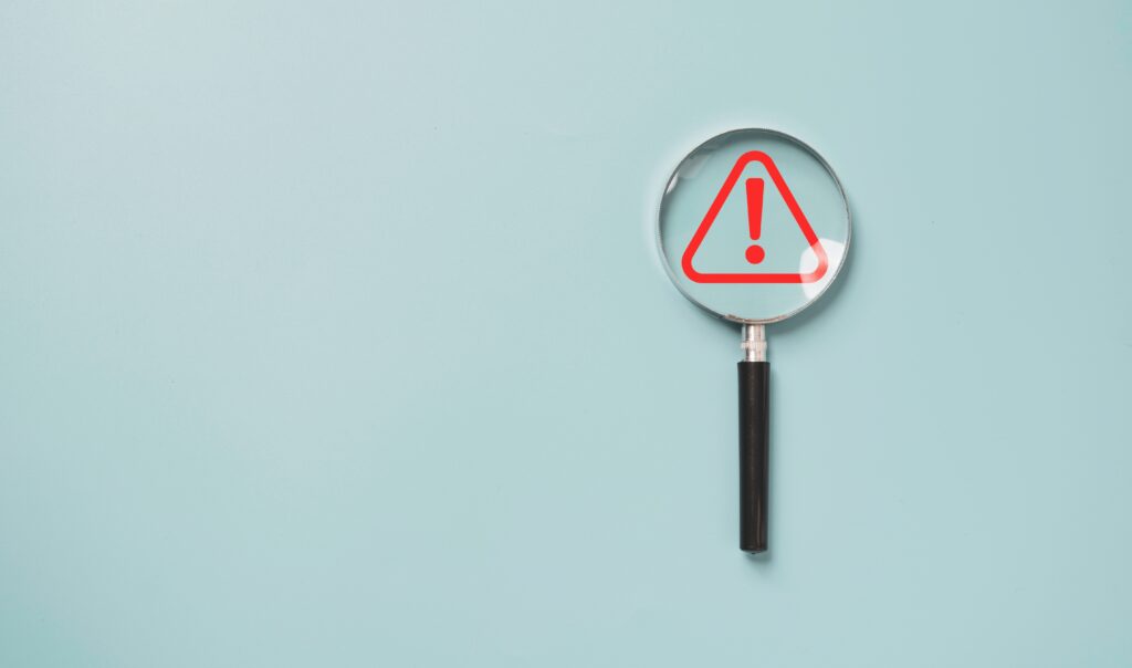 Magnifying glass highlights a red warning symbol, representing risks of hiring the wrong benefit plan auditor.