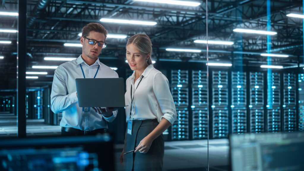 Two IT professionals review data on a laptop in a server room, emphasizing cyber security and accounting infrastructure.