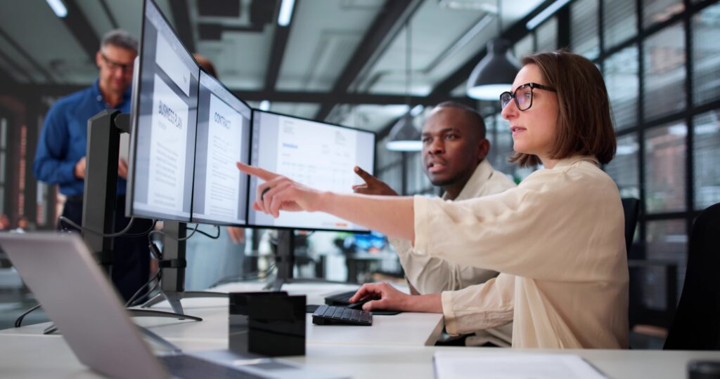 Team reviews documents on dual monitors in an office while discussing new pcaob auditing standards.