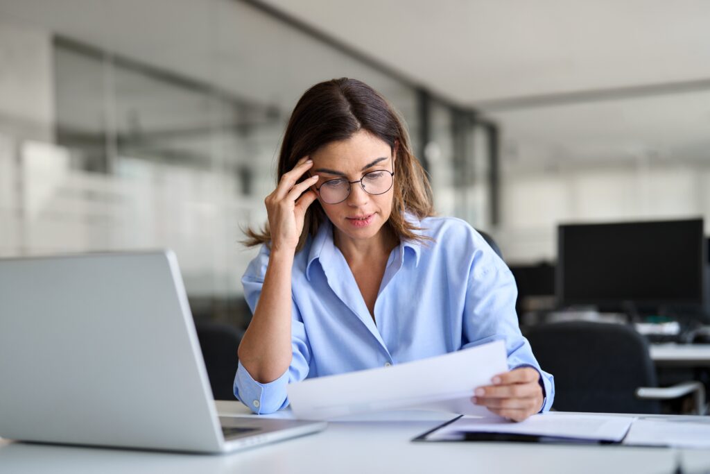 A business woman looking frustrated while 403 b audit paperwork ris eviewed at a desk with a laptop and folders in a quiet office